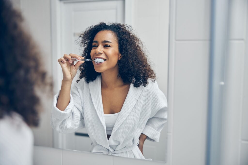 Woman in white bathrobe brushing teeth at bathroom mirror
